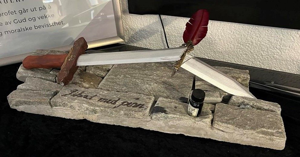 Et knekt sverd men en fjærpenn oppå er utstilt på et bord. Foran står det skrevet "Jihad med penn". Foto.