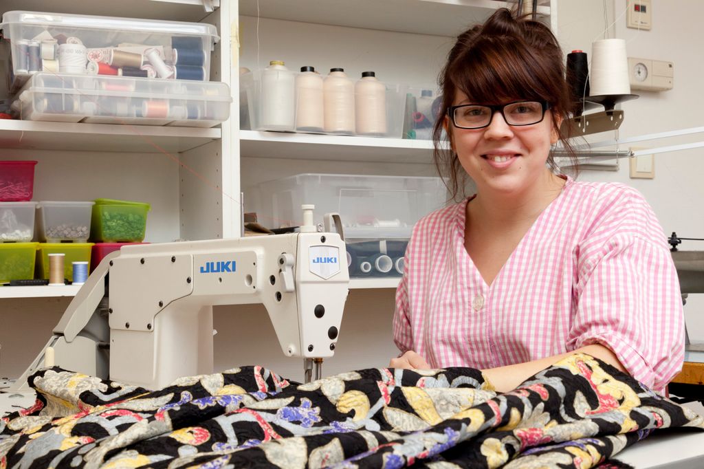 Fotografi av en smilende kvinne som sitter foran en symaskin og syr møbelstoff.
