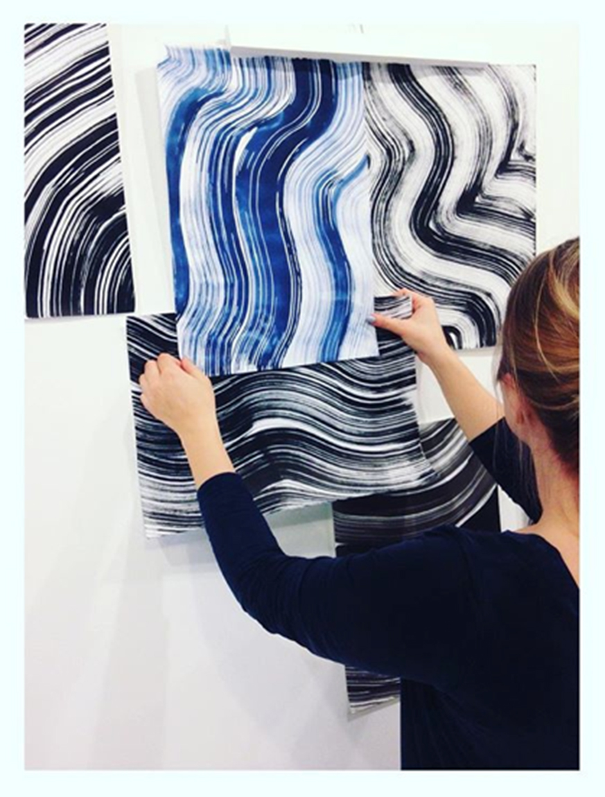 Designer vurderer ulike tekstiltrykk med bølgede linjer i blått, hvitt og svart. Hun holder dem opp mot veggen. Foto.