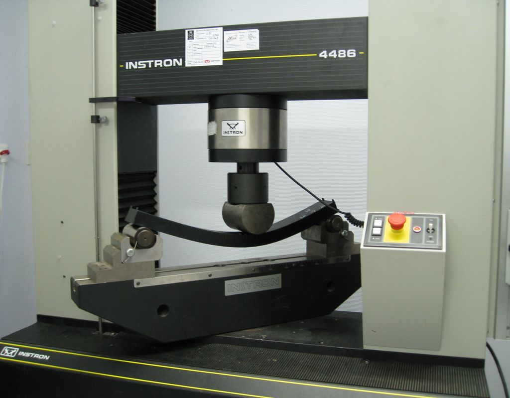 Maskin som presser et stempel ned på en metallplate som ligger på to støttebukker. Foto.