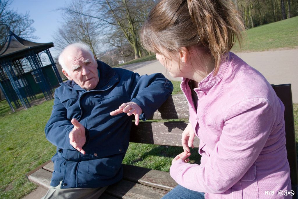Eldre mann i samtale med en kvinne, de sitter på en benk utendørs. Foto.