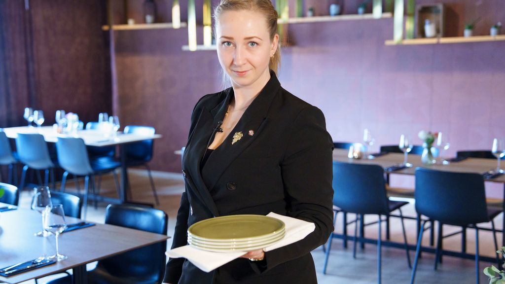 En servitør i en restaurant bærer en stabel med tallerkener i en hånd. Under tallerkenene har hun en håndserviett. Foto.