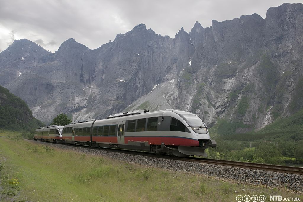 NSB-tog som kjører på togskinner i natur med grønne sletter og fjelltopper i bakgrunnen. Foto. 