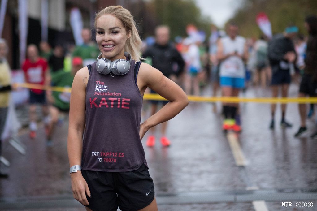 Woman, Katie Piper, in training gear. Behind her is a yellow ribbon accross the road, the background is blurred but we can see runners standing there. Photo.