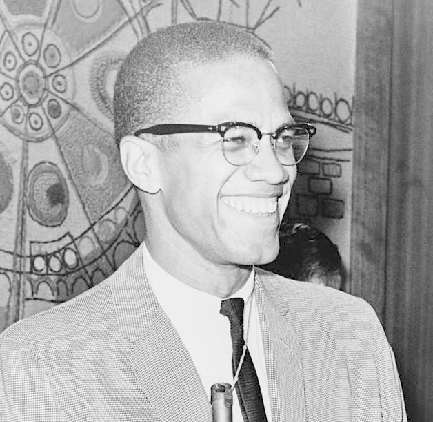 Photo: We see a Black man. he is wearing a suit and tie. he has glasses. He is smiling.