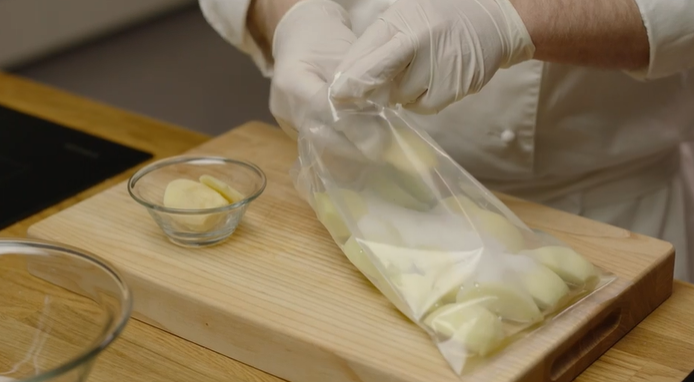 Nærbilde av hender med plasthansker som legger eplebåter i en pose. Foto.