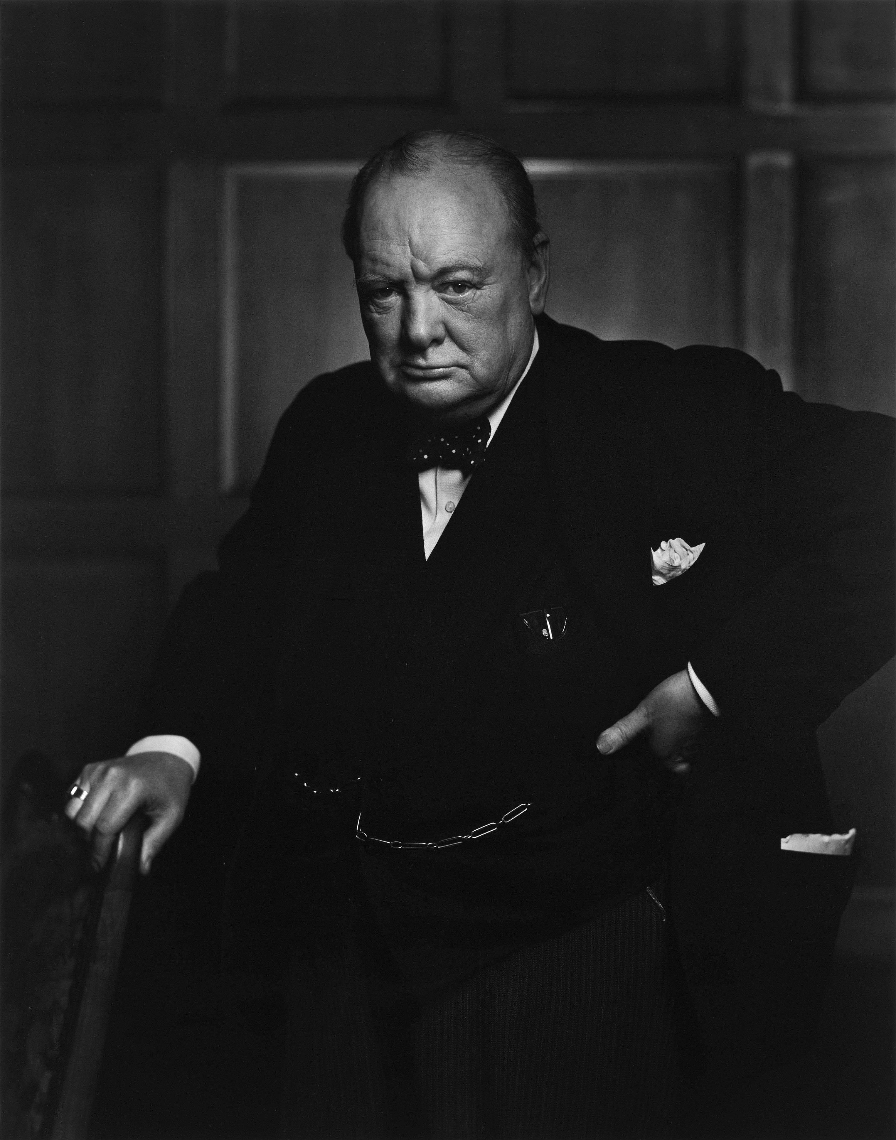 Portrait of Winston Churchill holding a cane. We see a stout, older man. He is wearing a dark jacket, bow tie, and a white shirt: He has a pocket handkerchief. He looks serious. Photo.