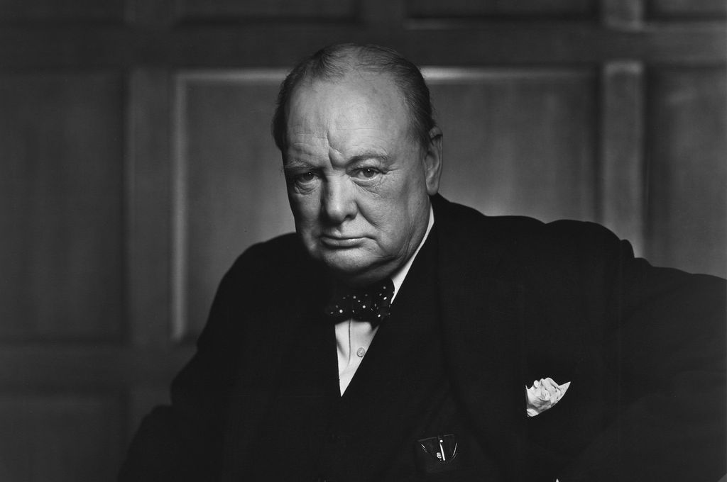 Portrait of Winston Churchill holding a cane. We see a stout, older man. He is wearing a dark jacket, bow tie, and a white shirt: He has a pocket handkerchief. He looks serious. Photo.