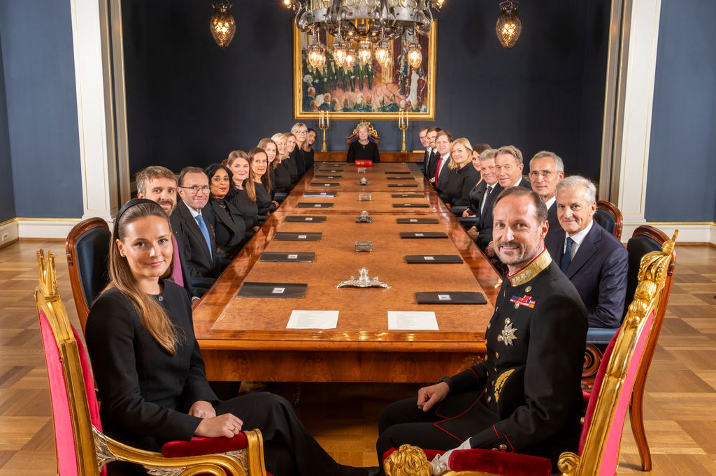 Ei gruppe menneske ved eit langbord ser høgtideleg i kamera. Alle er pent kledde og den næraste bærer mørk uniform. Biletet er av regjeringa Støre i statsråd med kong Haakon og kronprinsesse Ingrid Alexandra. Foto.