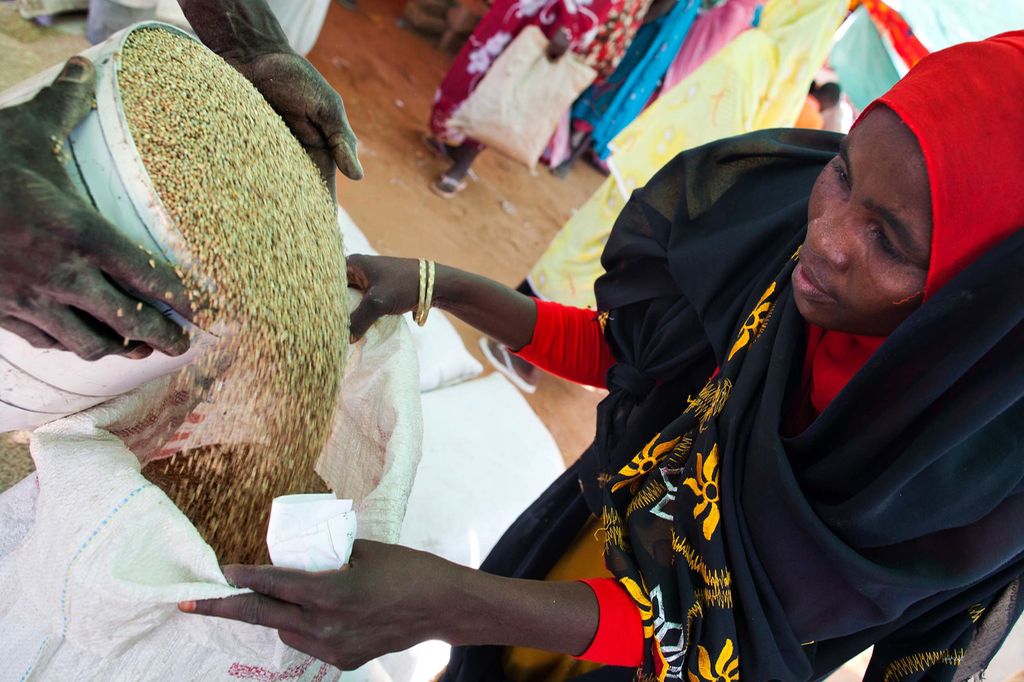 Black woman in red head scarf holds bag open as a bucket of grain is poured into it. Photo.