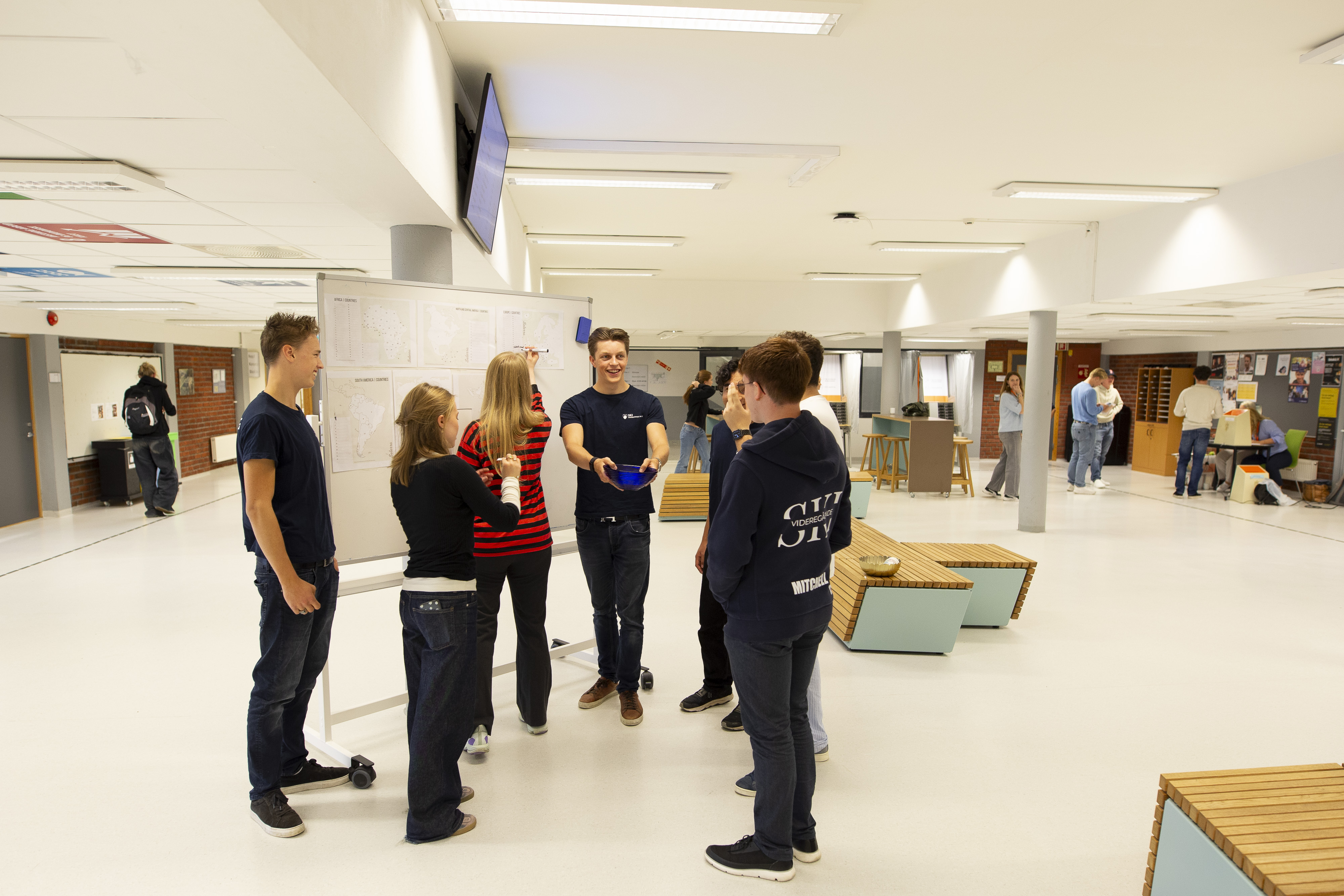 Sju elever står rundt ei tavle i aulaen på skolen. Foto.
