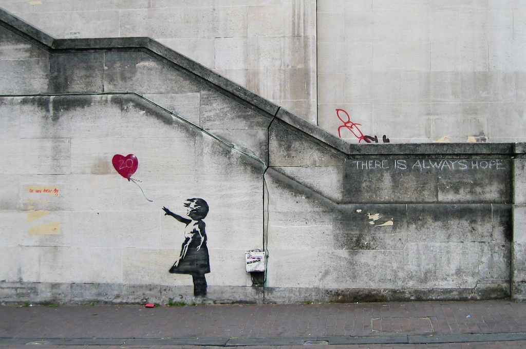 Graffiti av ei lita jente malt på en murvegg. Hun har vinden bakfra og en rød ballong formet som et hjerte er i ferd med å blåse bort fra henne. Litt til høyre for jenta, på samme murvegg, står det skrevet "There is always hope". Foto.