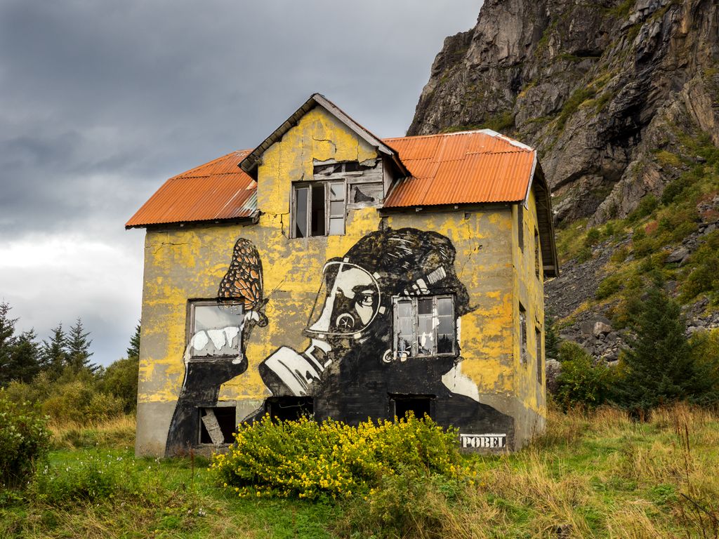 Graffiti på et falleferdig gult hus. En person med gassmaske ser på en stor sommerfugl som balanserer på pekefingeren hans. Foto.