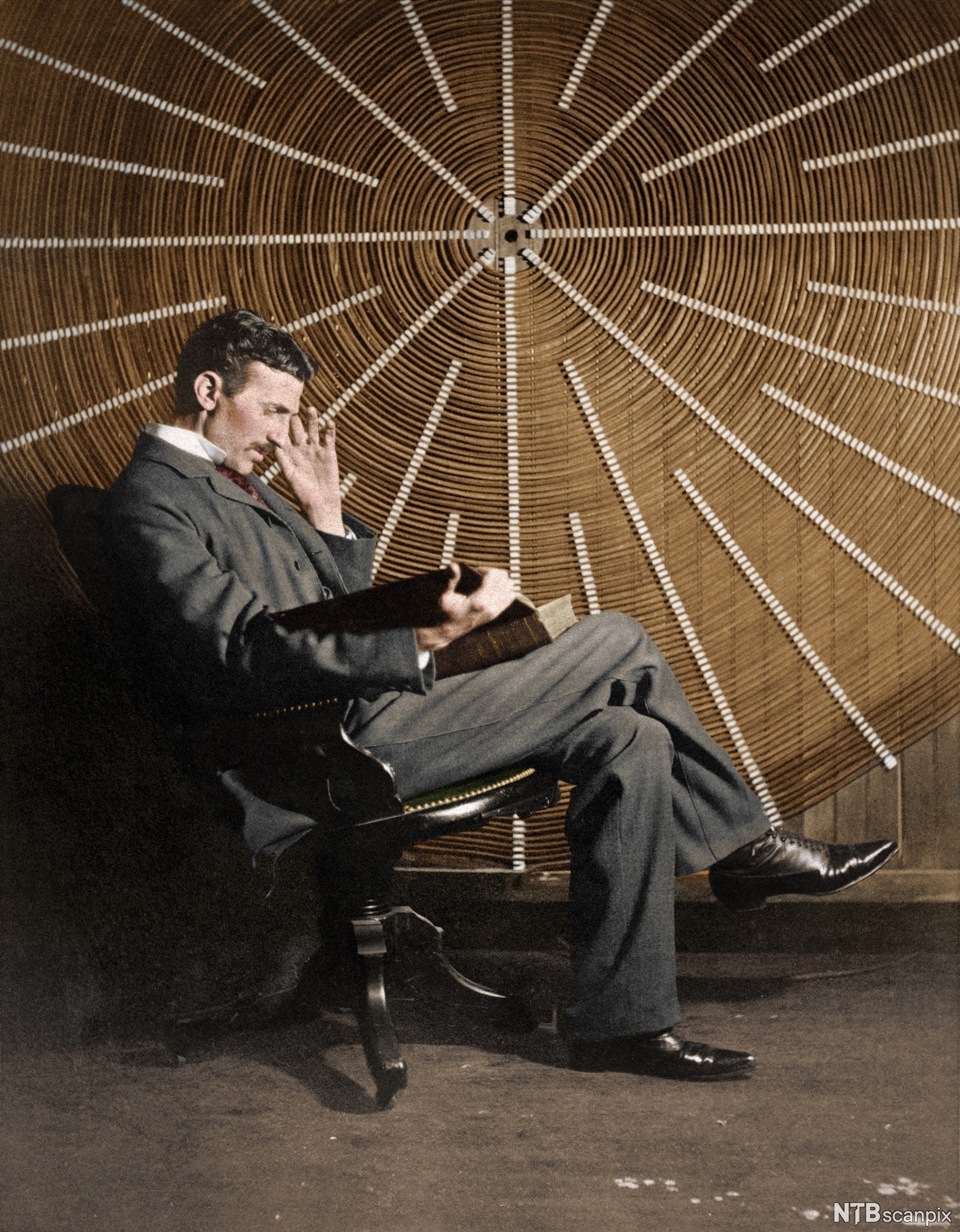 Photo: Nikola Tesla seated in front of a large coil. He is wearing a suit and black leather shoes. 