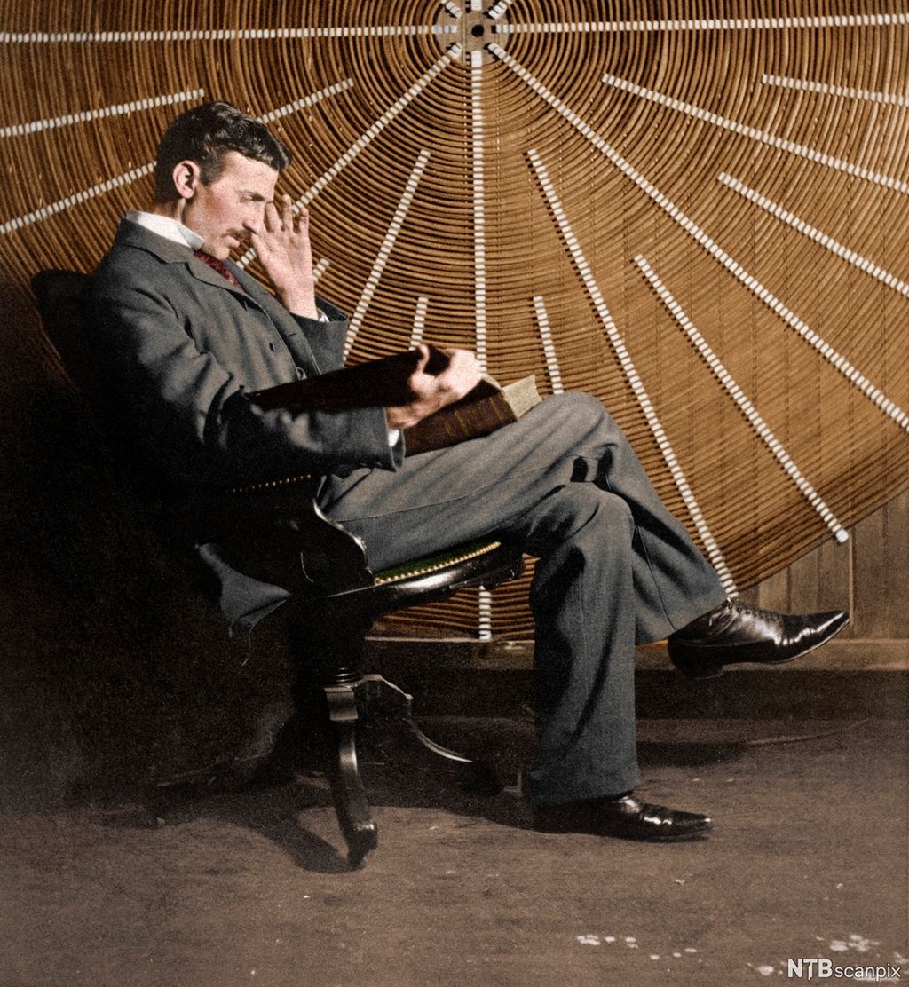 Photo: Nikola Tesla seated in front of a large coil. He is wearing a suit and black leather shoes.