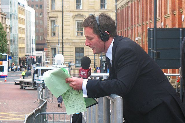 En dresskledd radiojournalist står lent mot et avsperringsgjerde i metall. Han har hodetelefoner på og leser i en håndholdt mikrofon fra en notatblokk. Foto.