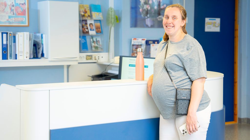 Gravid kvinne står ved ein skranke. Ho ser inn i kamera og smiler. Foto.