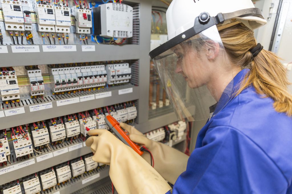 En ung elektriker på jobb. Hun har isolerhansker, hjelm, visir og kjeledress. Foto.