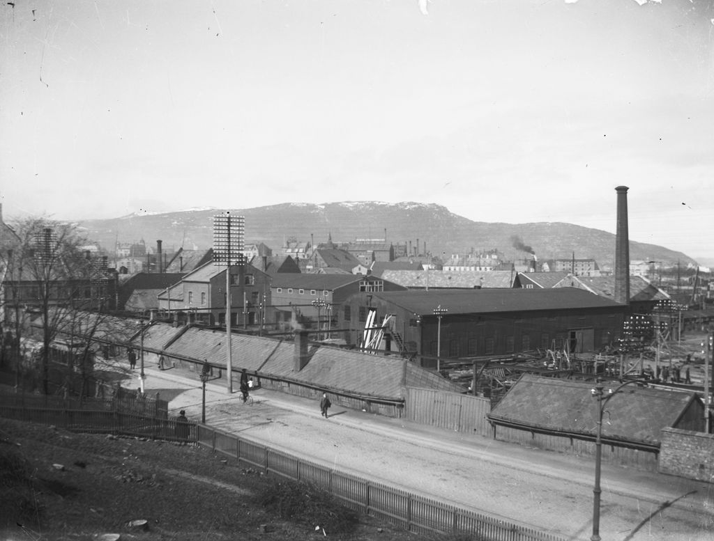 Ulike typer bygninger nedenfor en vei der vi ser noen få gående. Det er bygninger på både tvers og langs ved siden av veien. Dette er reperbanen og Trondhjems Mekaniske Verksted. Foto.