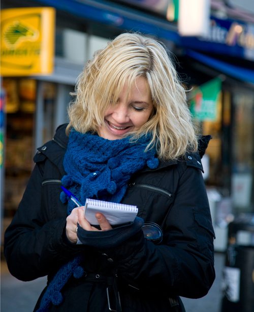 Ei kvinne med jakke og skjerf står i ei gate og noterer på ei notisblokk. Foto.