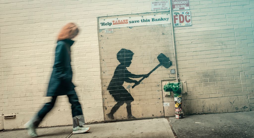 The street art graffiti, called ‘Hammer Boy’, is a piece by Banksy. It depicts a child with a hammer, poised to smash a fire hydrant. Above the artwork, the text reads, ‘Help ZABAR save this Banksy.’ A blurred person can be seen walking past the mural. Photo.