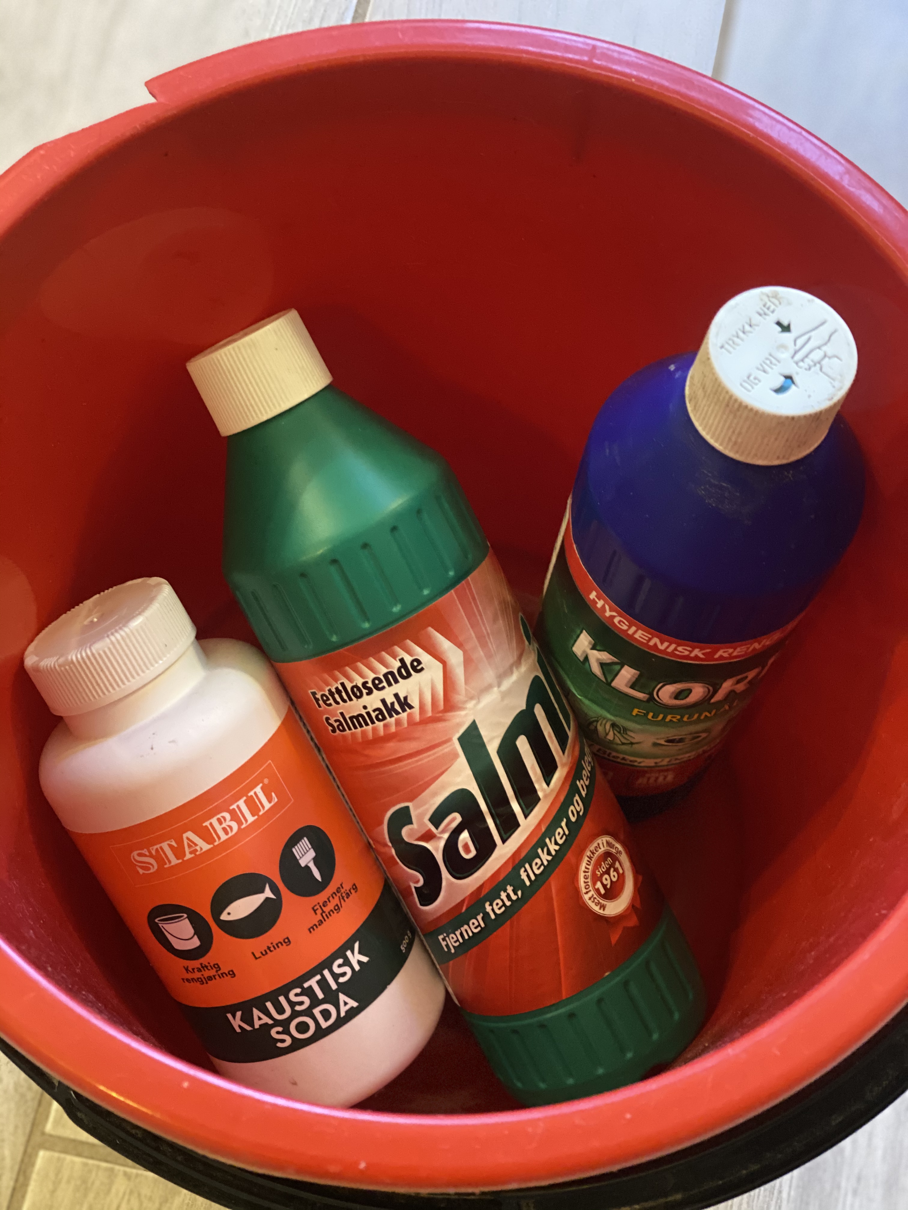 En rød bøtte med en flaske kaustisk soda, en flaske salmiakk og en flaske klorin. Foto.
