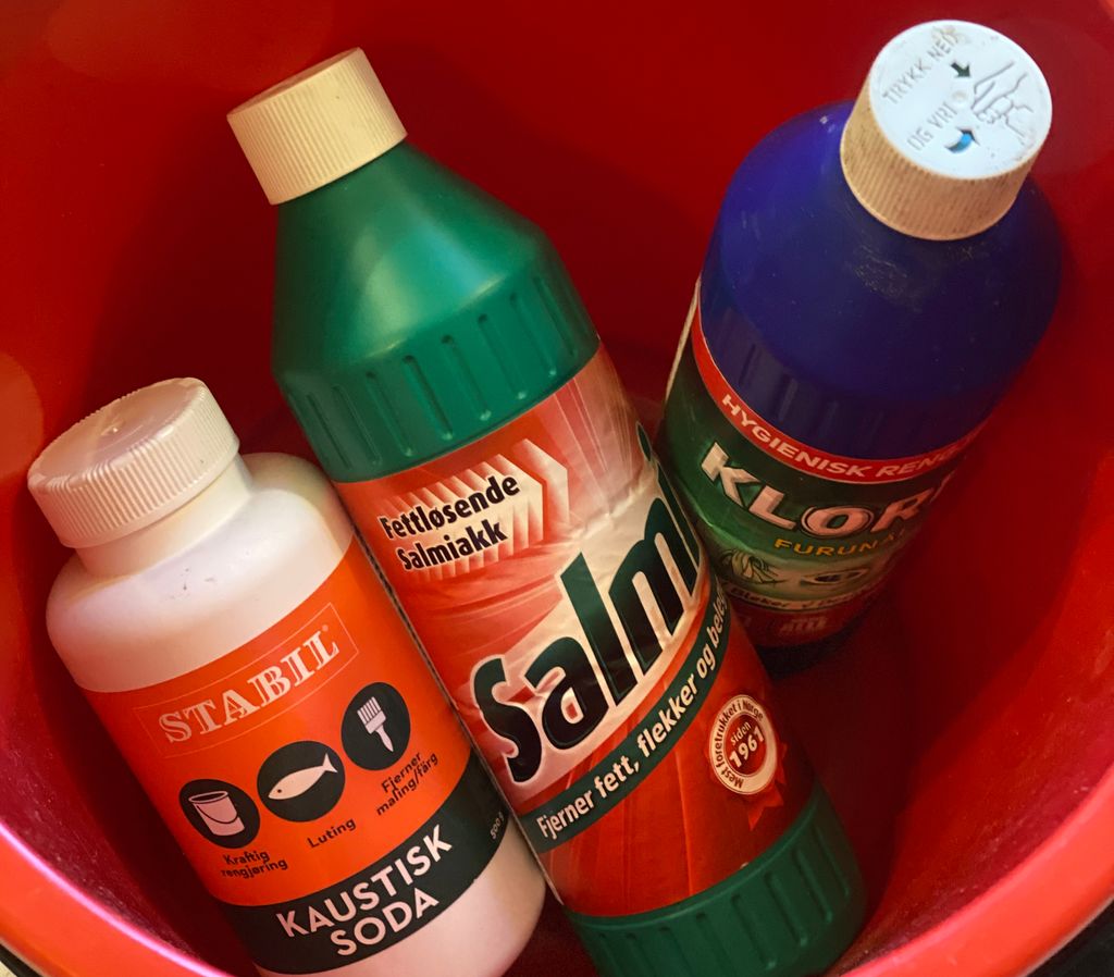 En rød bøtte med en flaske kaustisk soda, en flaske salmiakk og en flaske klorin. Foto.