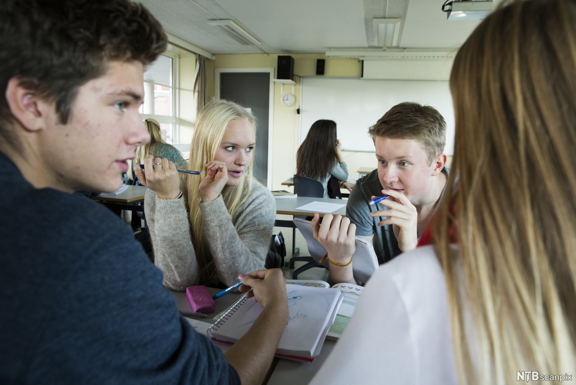 Foto av fire ungdommer som diskuterer rundt en pult i et klasserom.
