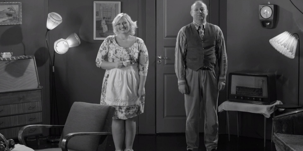 Mann og dame står og smiler mot kameraet. Bildet er i svart-hvitt, og klærne og dekoren er fra 1960-tallet.
