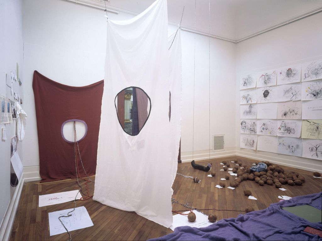 Installasjon som inneholder tegninger og utklipp på veggene, tekstiler med sirkelformede hull henger ned fra taket, og på gulvet ligger tekstiler, tegninger og kokosnøtter. Foto.
