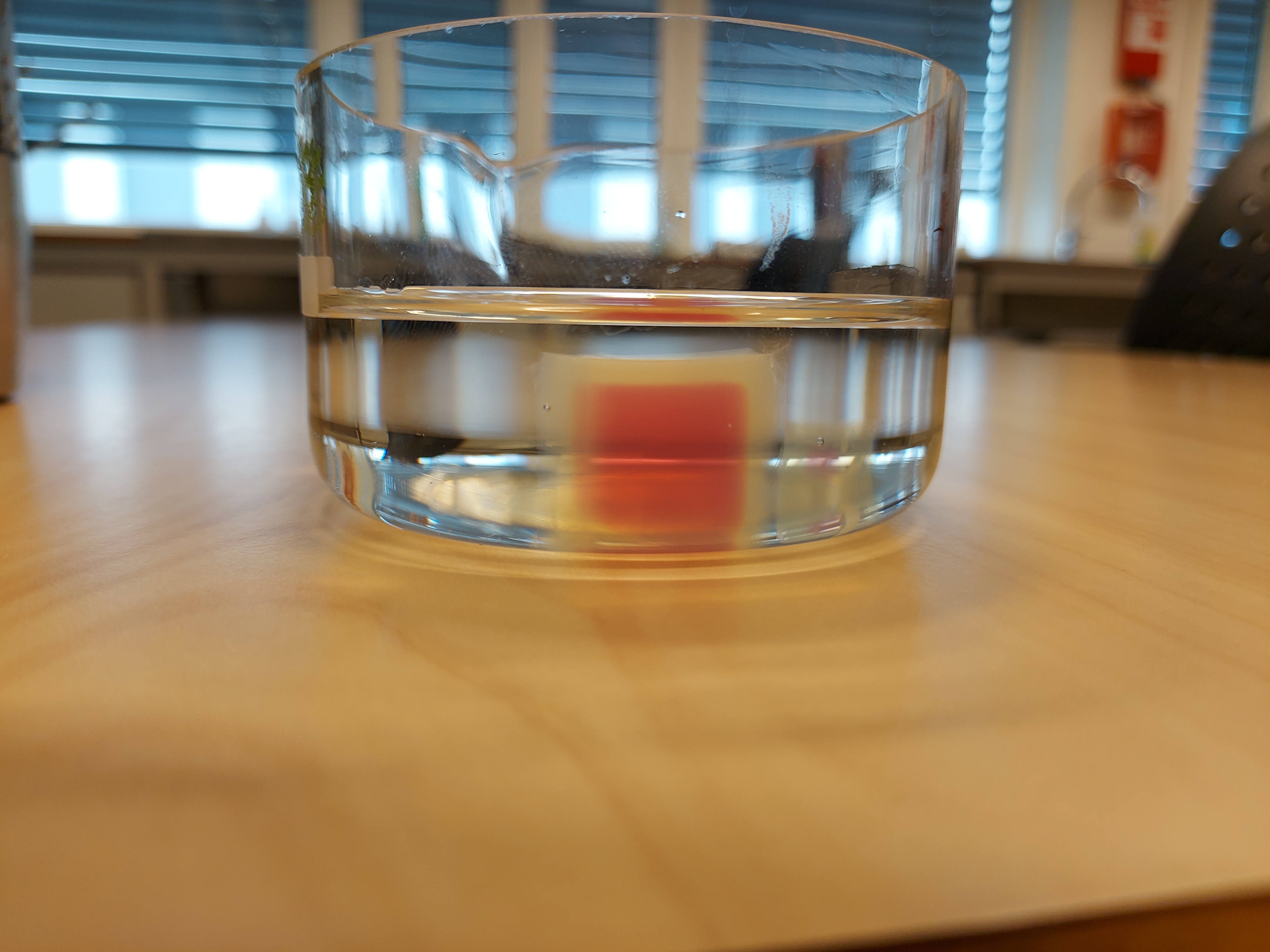 Geléaktig terning med rødt felt ligger i væske i et glass. Foto.