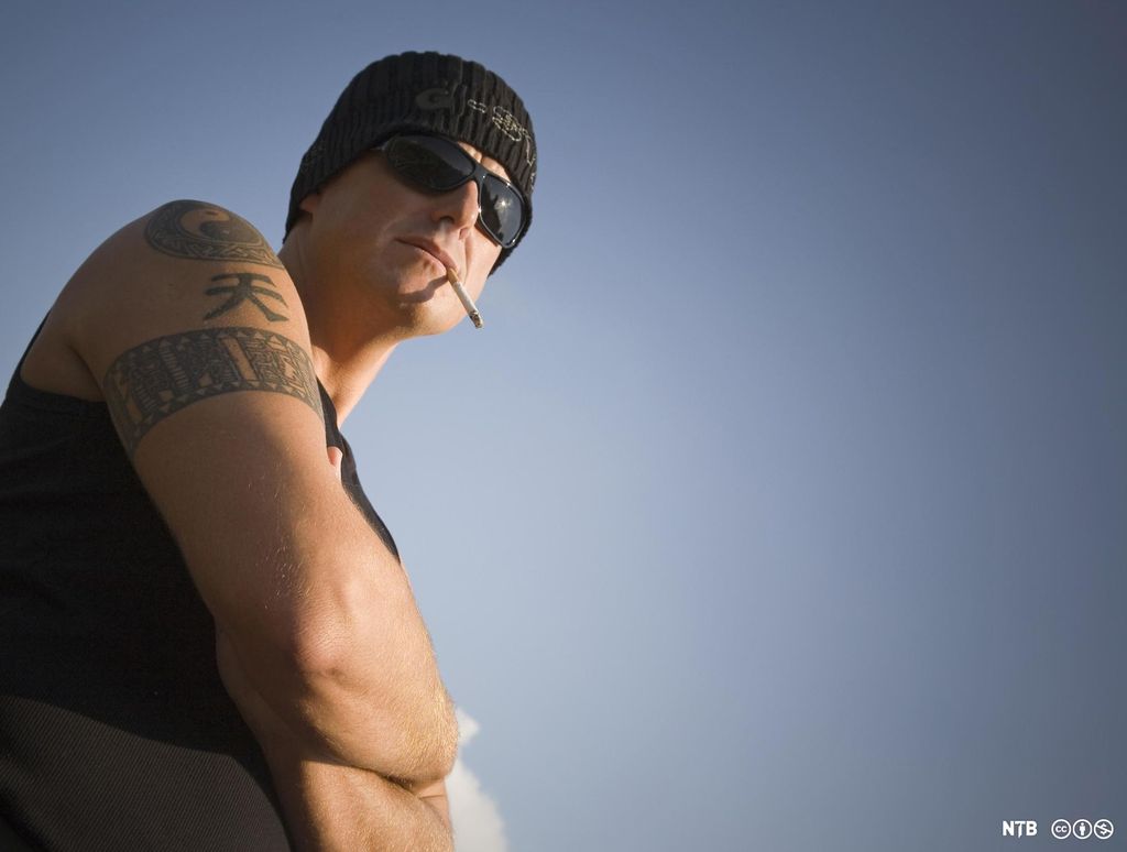 En mann med tatoveringer, lue, solbriller og sigarett i munnviken ser mot kamera. Foto.