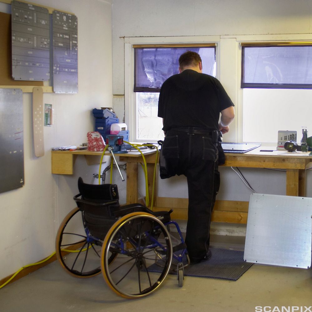Rullestolbruker som arbeider ved en arbeidsbenk. Personen har kun ett bein, men står oppreist ved pulten. Rullestolen er bak ham. Foto.