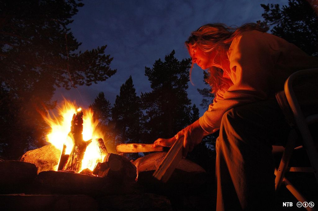 Ei kvinne sitter ved et bål i en skog. Hun legger ved på bålet. Det er mørkt rundt henne. Foto.