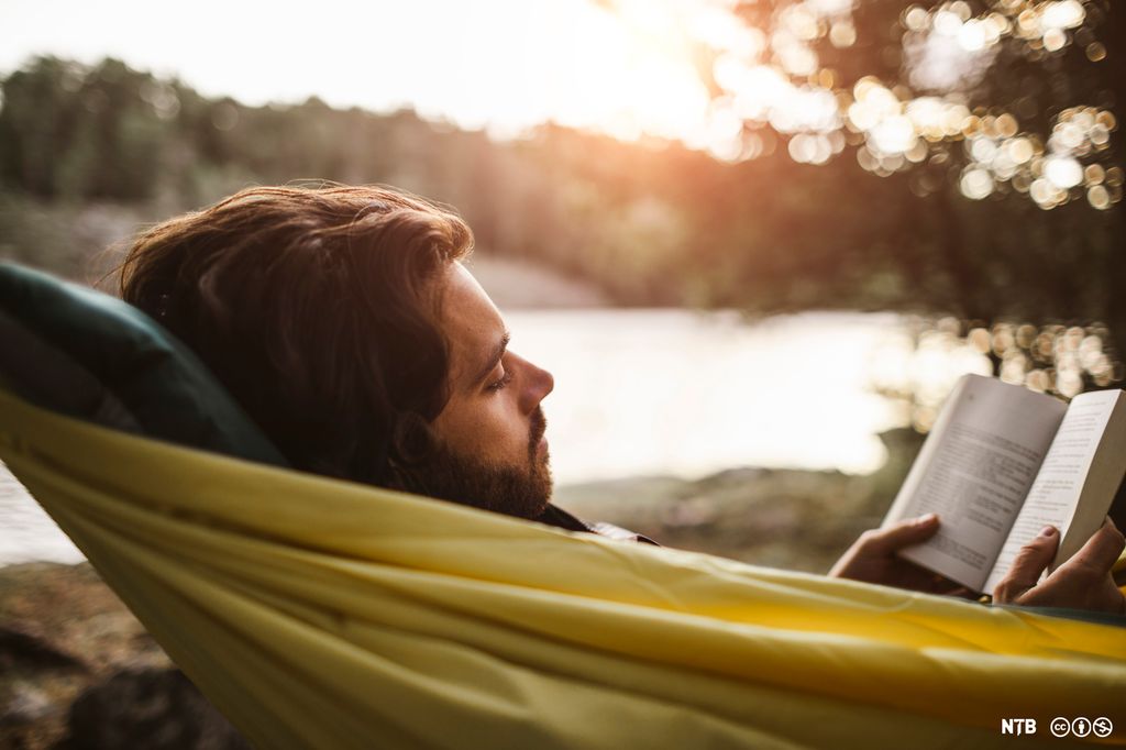 Ung mann ligger i hengekøye og leser bok. Foto.
