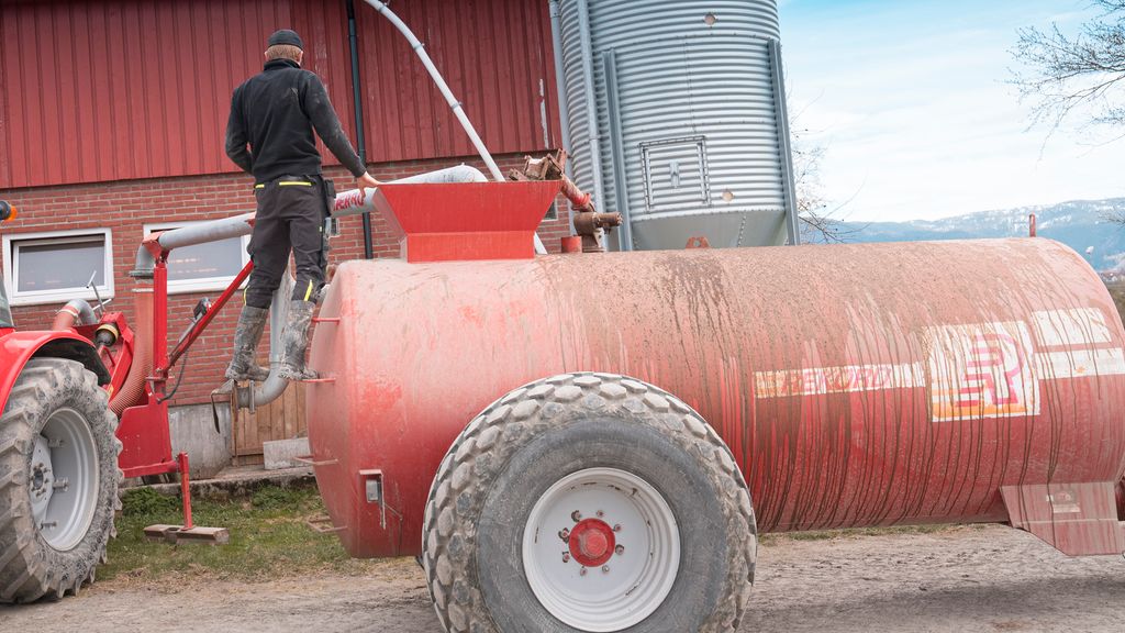 En bonde står bakpå ei gjødselvogn og følger med. En slange kommer fra et fjøs i bakgrunnen og opp i gjødselvogna. Foto.