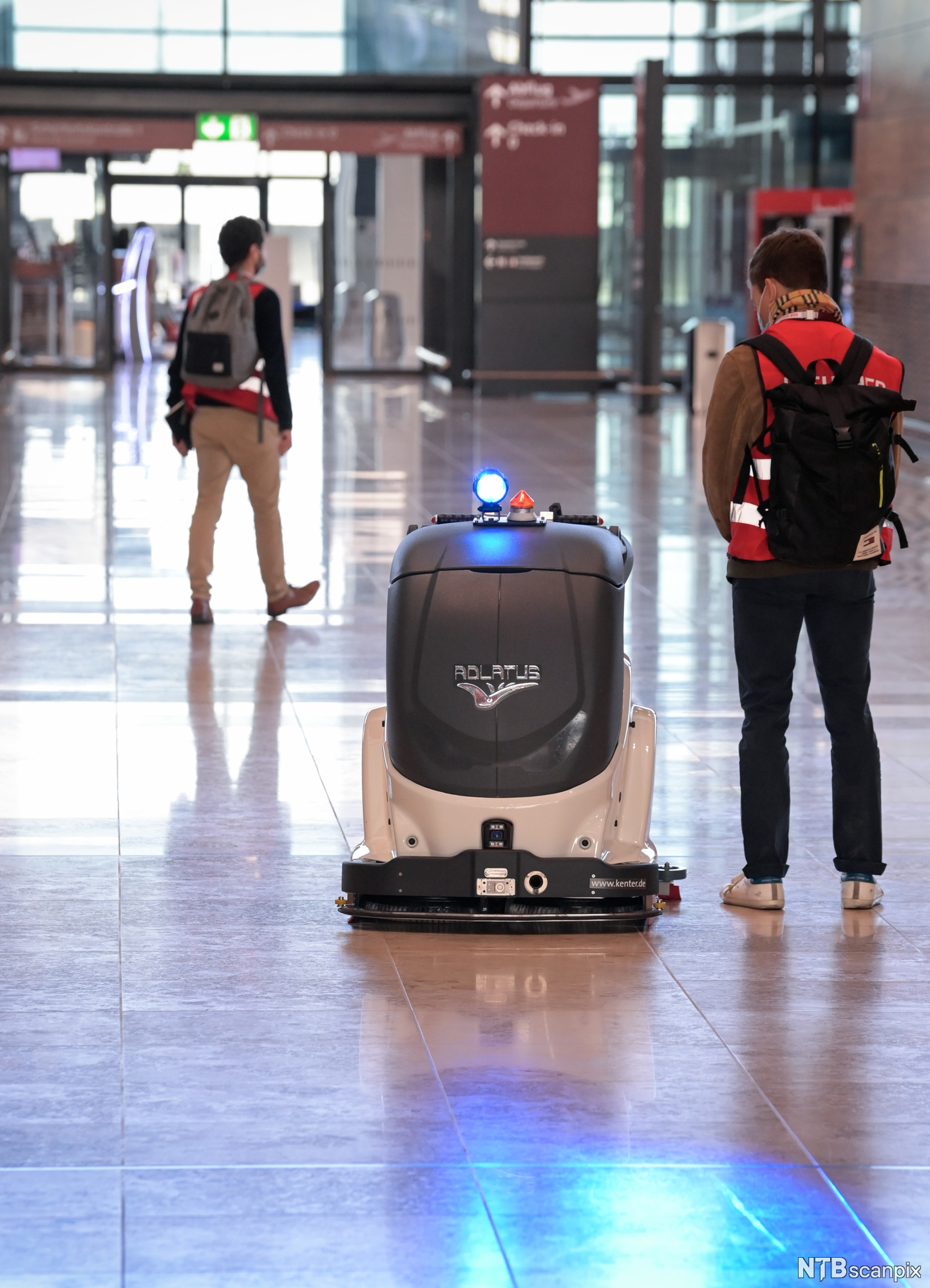 En hvit og grå vaskerobot i en hall på en flyplass. Et menneske står ved siden av. Foto.