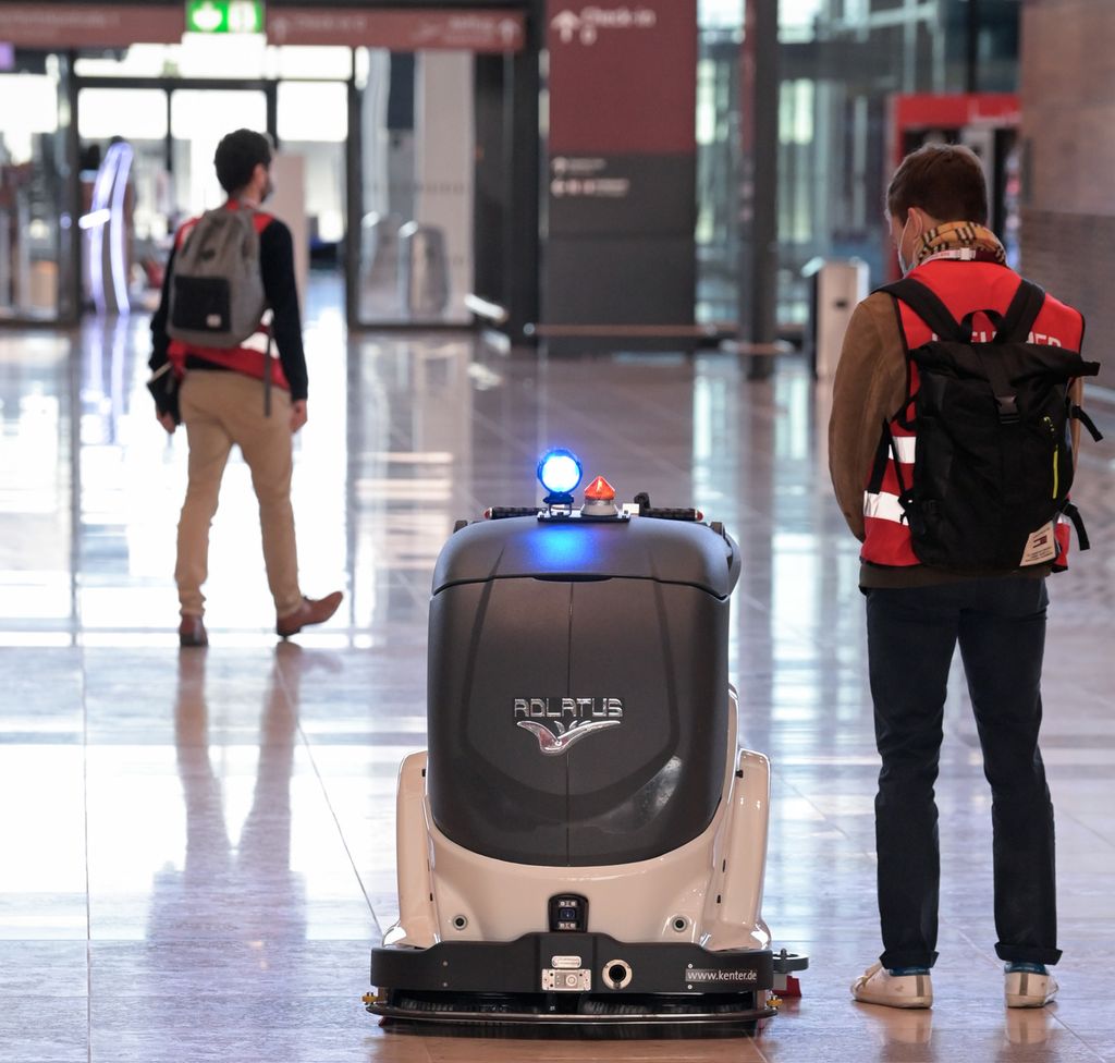 En hvit og grå vaskerobot i en hall på en flyplass. Et menneske står ved siden av. Foto.