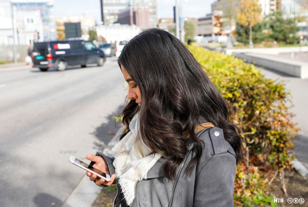 Ei jente på 17–18 år med langt, mørkt hår står ved en trafikkert vei i en by. Hun ser ned på mobilen som hun holder i handa. Ansiktsuttrykket er nokså alvorlig. Jenta har på seg ei grå skinnjakke og et hvitt skjerf. Foto.