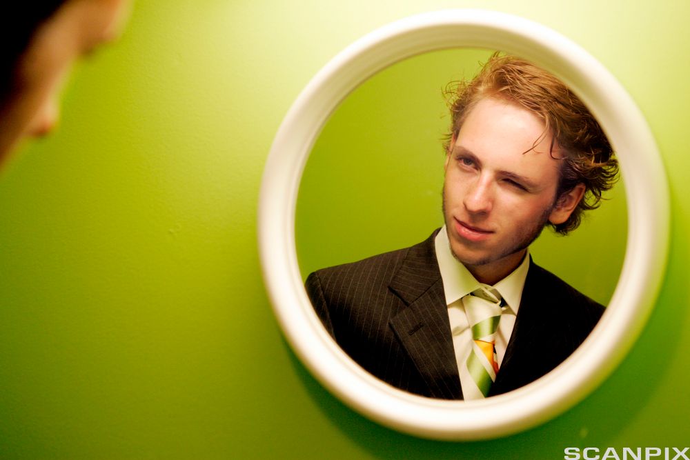 En selvsikker, ung mann med slips ser på seg selv i et speil mens han blunker med et øye og smiler lurt. Foto. 