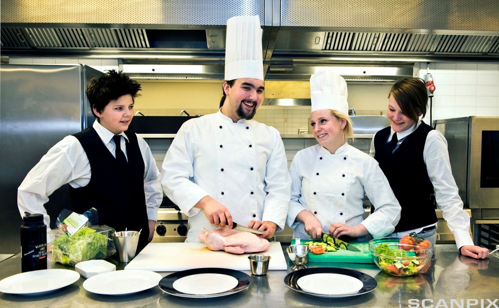 En kokk skjærer opp en kylling og en kokkelærling hakker grønnsaker. To servitørlærlinger står på hver sin side av kokkene og observerer. Foto.