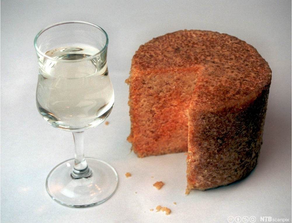 En rund, rødbrun gammelost hvor et stykke er skåret ut. Ved siden står vinglass fylt med blank drikke. Foto.