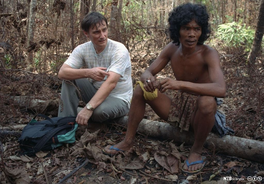 En mann i vestlige klær og en mann med lendeklede og flippflopper sitter sammen på en trestamme i skogen.. Foto. 