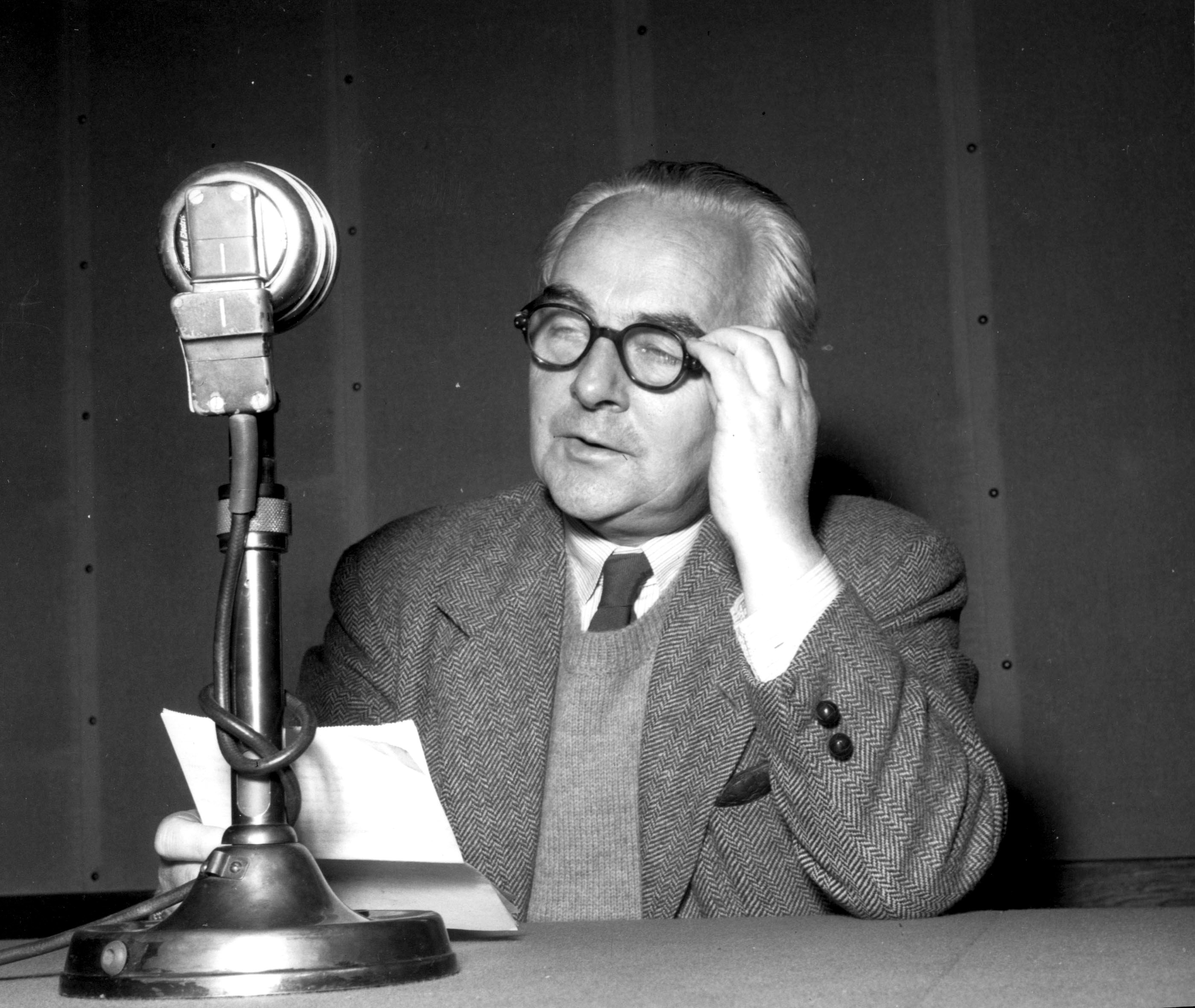 Mann med strikket vest, tweedjakke og tykke brilleinnfatninger holder et ark og sitter ved en bordmikrofon. Foto.