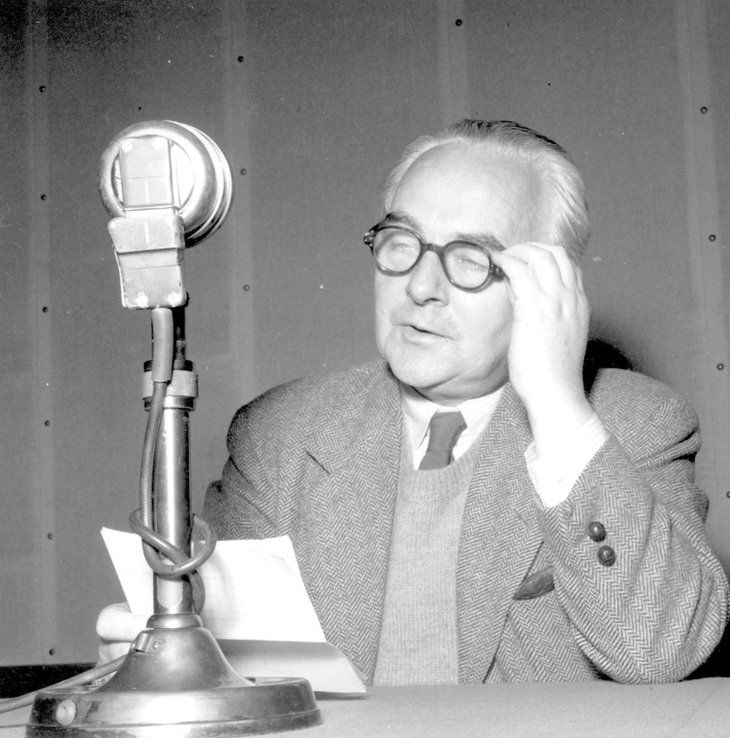 Mann med strikket vest, tweedjakke og tykke brilleinnfatninger holder et ark og sitter ved en bordmikrofon. Foto.