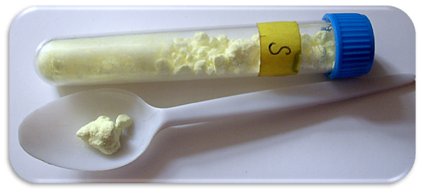 Eit reagensrør og ei plastskei med lysegult pulver. Reagensrøret er merka med S for svovel. Foto.