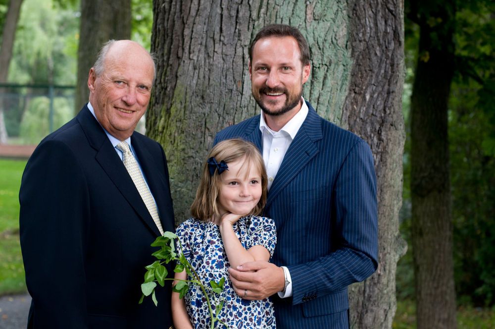 Kong Harald og kronprins Haakon i mørke dresser. Mellom dem står prinsesse Ingrid Alexandra i en prikkete kjole. Hun er fem år på bildet. De er fotografert fra hoftene og opp. Foto.