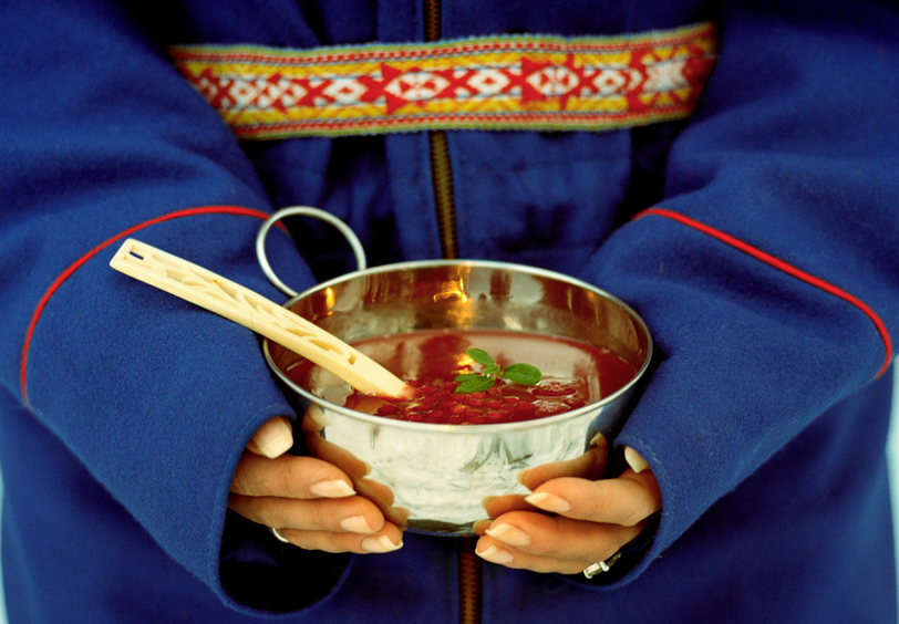 Kvinne i samekofte holder skål med tyttebærsuppe. Foto.
