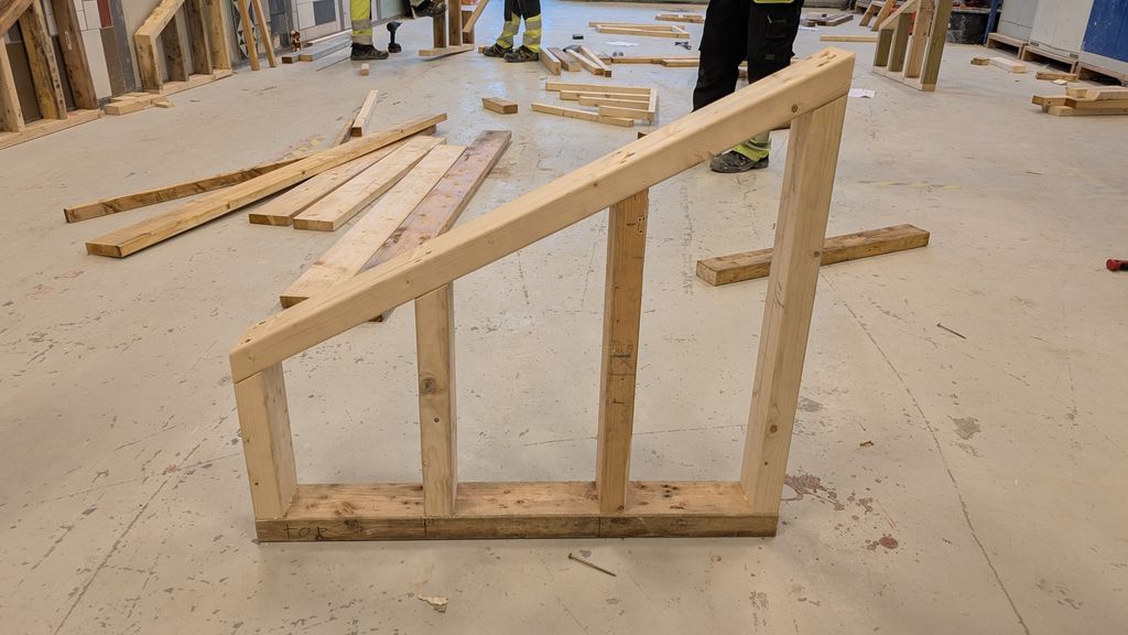 En ferdigbygget modul i reisverk stående i et verksted for byggfag. Modulen består av en bunnsvill og fire stendere med ulik lengde slik at toppsvillen går på skrå. Foto.