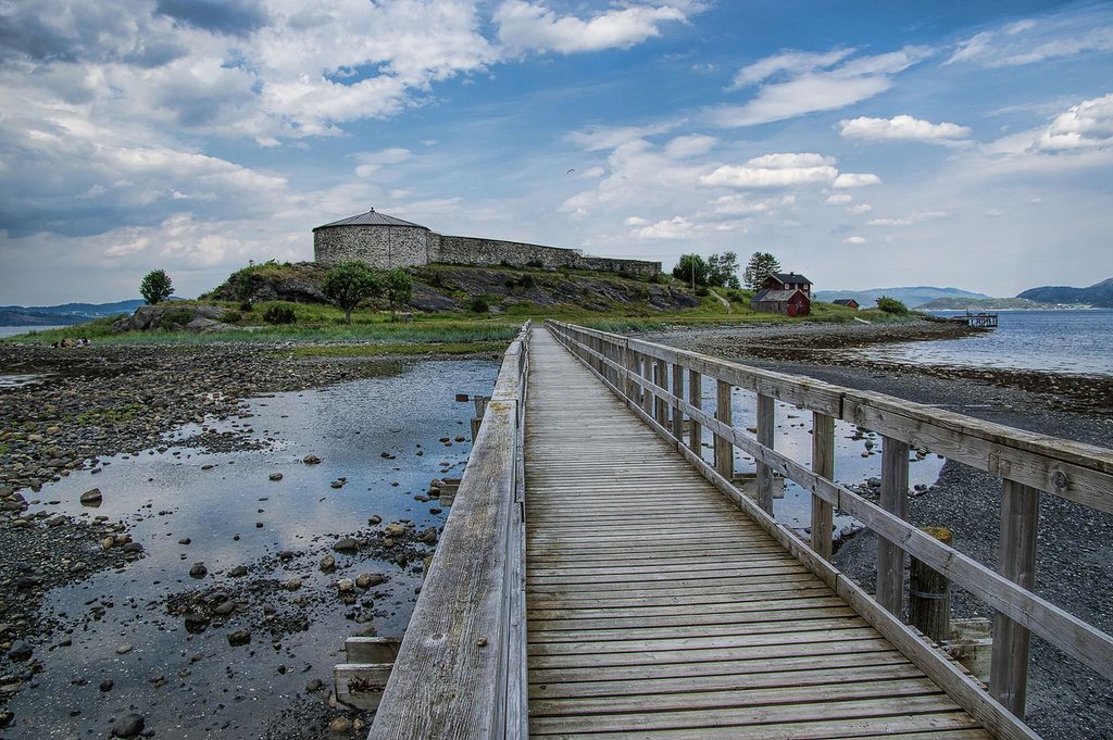 I forgrunnen ser vi trebroen som forbinder fastlandet med holmen der Steinvikholm slott ligger. I bakgrunnen ser vi steinfestningen øverst på den grønne holmen, omgitt av Åsenfjorden. Foto. 